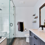 Modern bathroom with glass shower, double vanity with white countertop, large mirror, shelves with decor, vases with dried flowers, and a mix of black and white fixtures. Natural light enters through large windows.