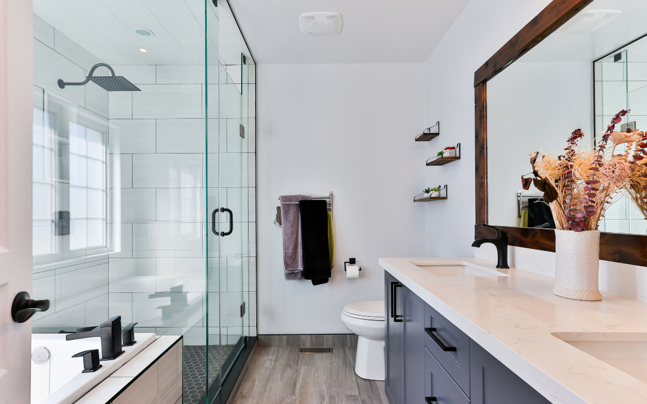 Modern bathroom with glass shower, double vanity with white countertop, large mirror, shelves with decor, vases with dried flowers, and a mix of black and white fixtures. Natural light enters through large windows.