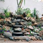 A small garden waterfall flows over stone steps surrounded by rocks, colorful flowers, and green plants, with a white wall and additional plants in the background.