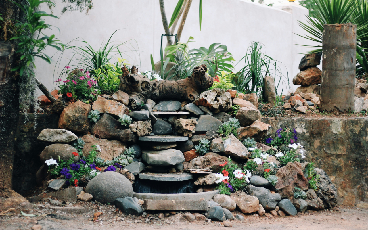 A small garden waterfall flows over stone steps surrounded by rocks, colorful flowers, and green plants, with a white wall and additional plants in the background.