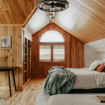 A cozy attic bedroom with wood-paneled walls and ceiling, a double bed with blankets and pillows, a small desk with a chair and vase, a window letting in natural light, and a coffee mug on a side table.