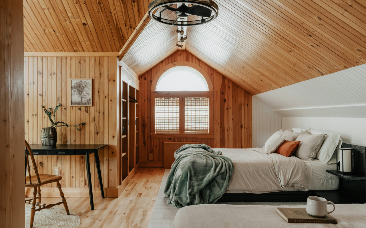A cozy attic bedroom with wood-paneled walls and ceiling, a double bed with blankets and pillows, a small desk with a chair and vase, a window letting in natural light, and a coffee mug on a side table.